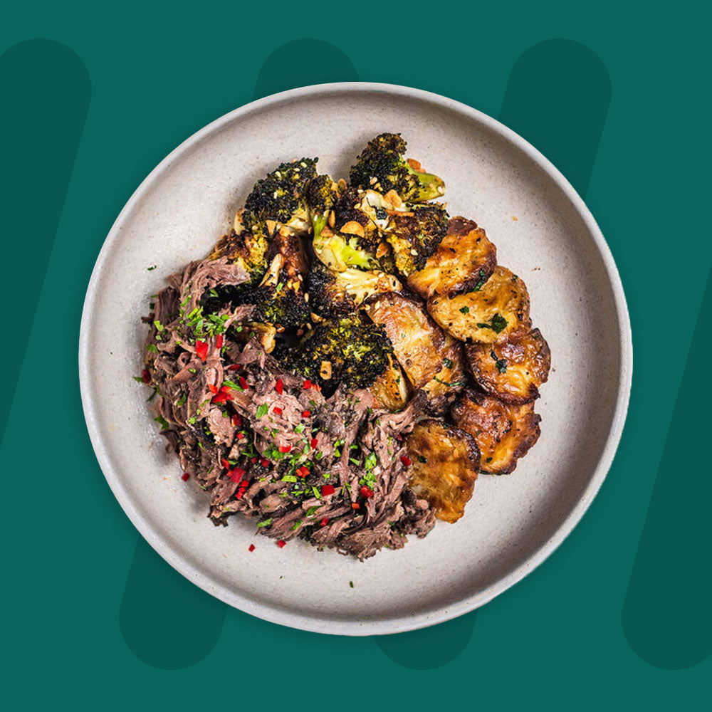 "A bowl featuring smoked pulled beef garnished with fresh herbs and red chili, garlic and herb crushed new potatoes, and charred broccoli with crispy garlic and sesame, set against a green background