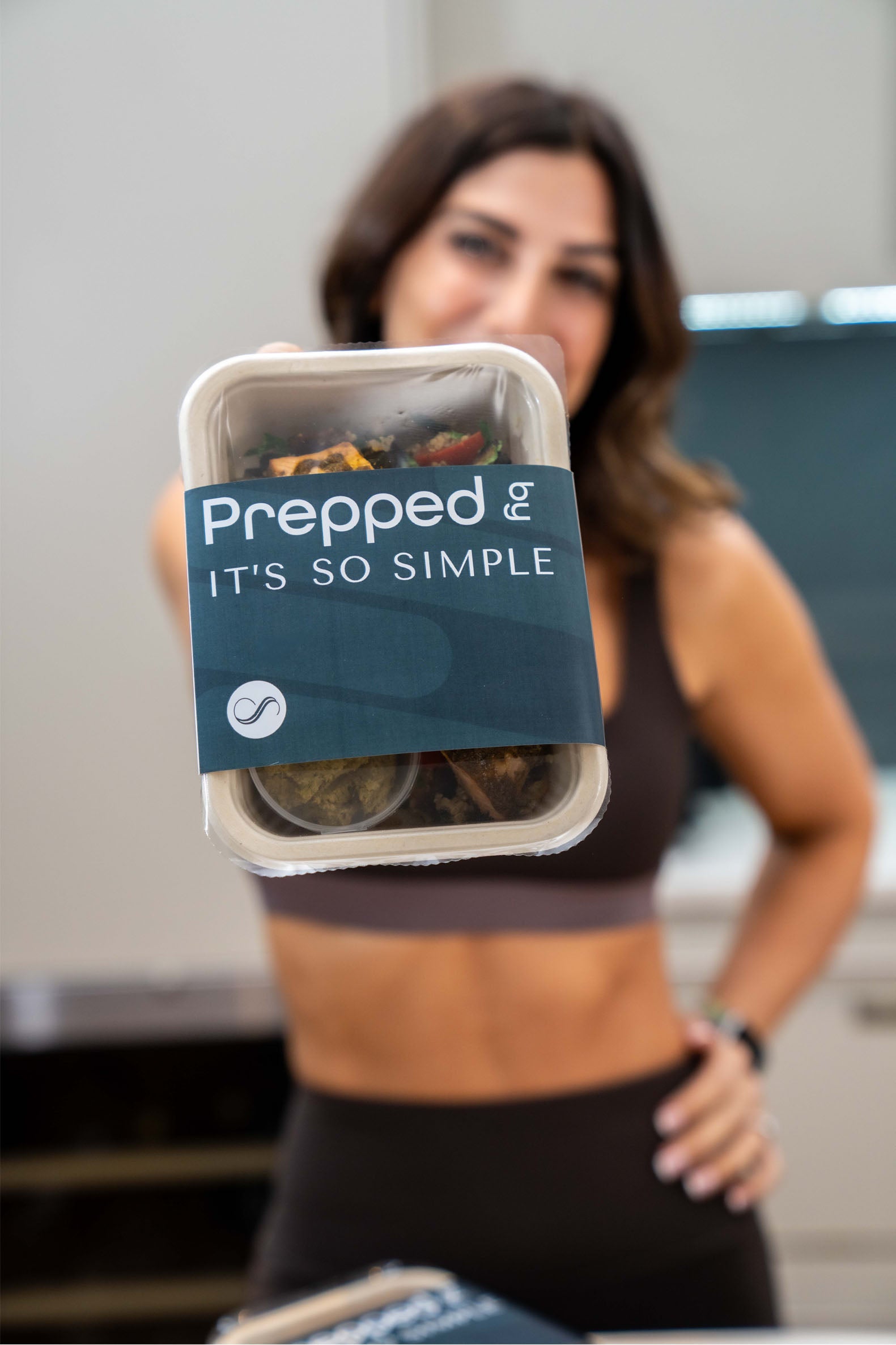 A woman in a cropped athletic outfit holds a pre-packaged meal container labeled 'Prepped - It's So Simple' prominently toward the camera. The container reveals a healthy meal with visible ingredients such as vegetables. The blurred background showcases a clean, modern kitchen.