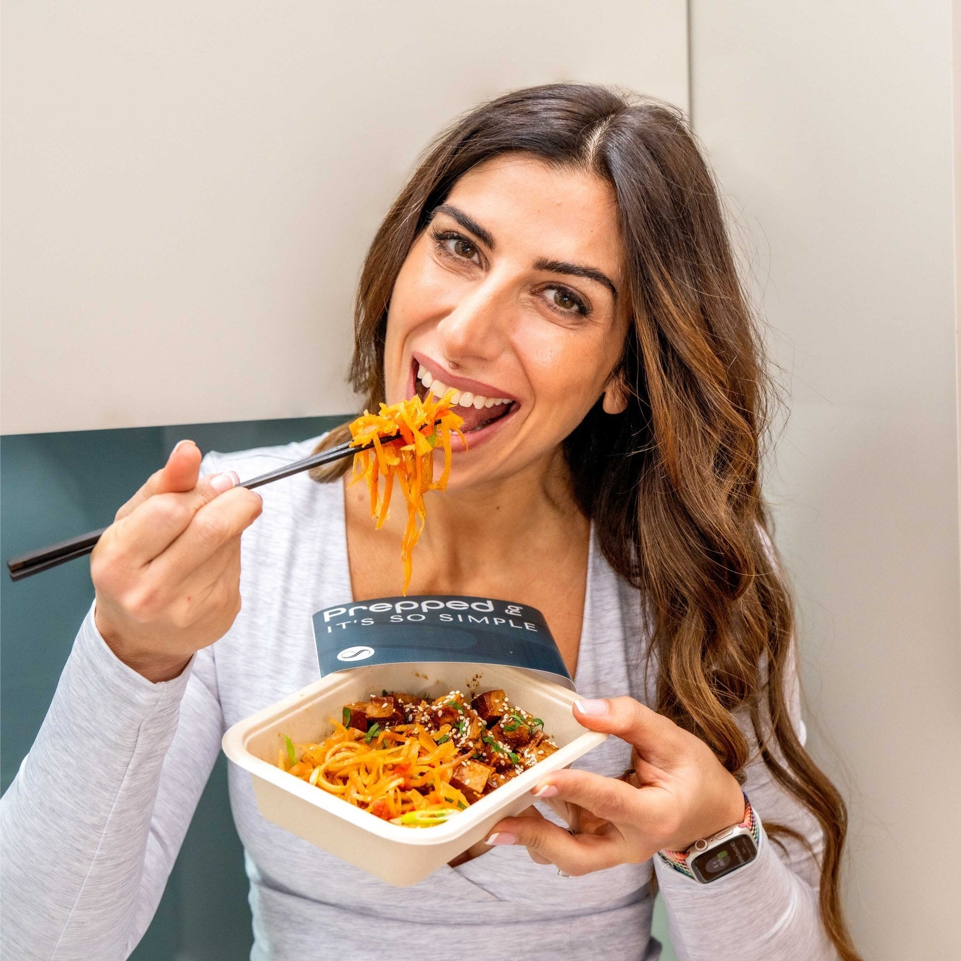 Rachael Sacerdoti, It's so Simple,A smiling woman with long wavy hair, wearing a light grey top and a smartwatch, holds a 'Prepped - It's So Simple' meal container. She uses chopsticks to lift a bite of vibrant orange noodles mixed with vegetables and garnished with sesame seeds and herbs. The background features a clean, minimal kitchen setup