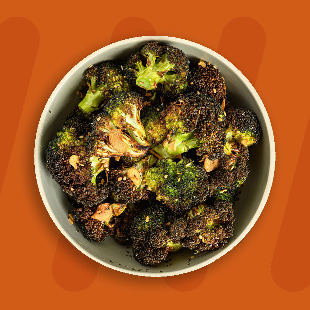 A bowl of charred broccoli florets, roasted to perfection with a crispy texture, garnished with toasted garlic slices and sesame seeds, set against an orange background.