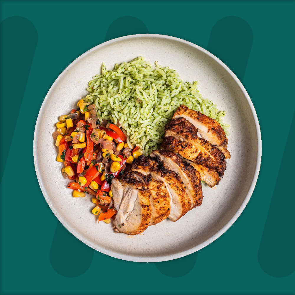 A plate featuring grilled, sliced chicken breast, accompanied by vibrant green herb rice and a colorful mix of roasted corn, red peppers, and onions, set against a green background.