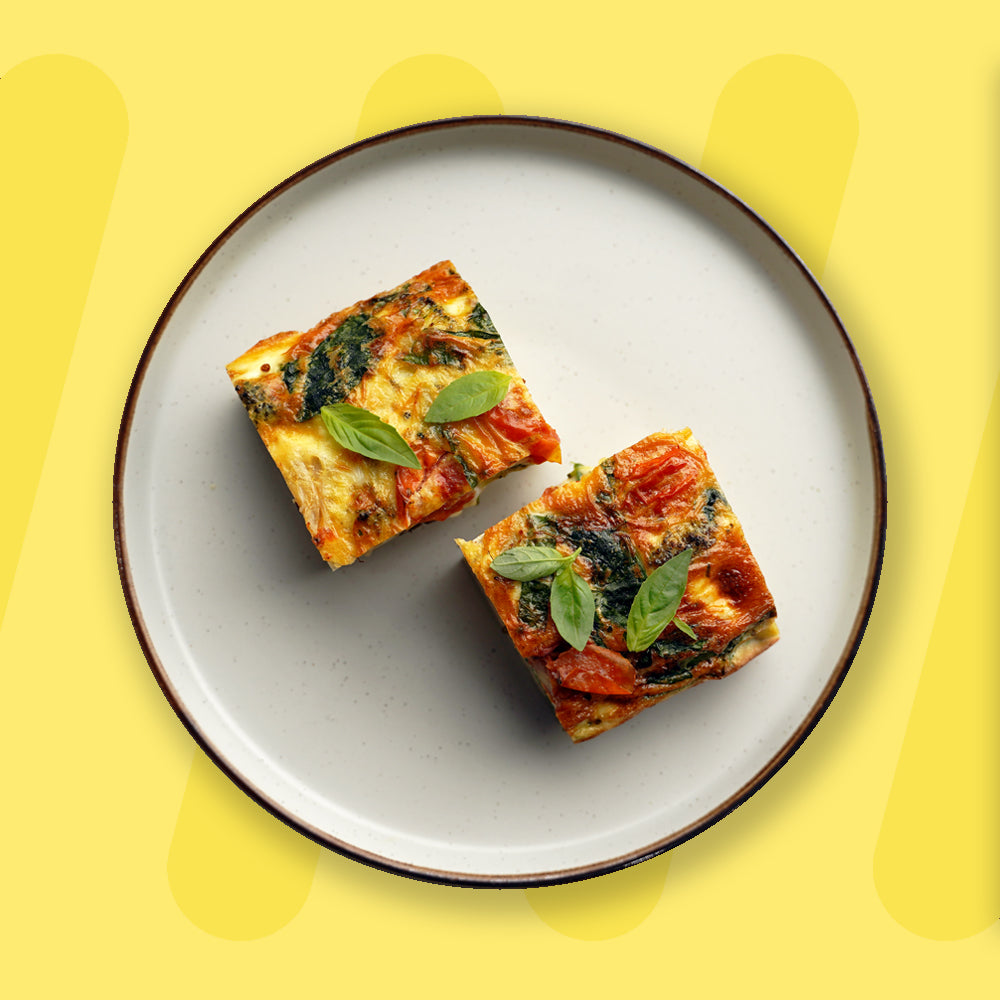 A plate with two square slices of vibrant vegetable frittata, featuring golden-brown edges, fresh spinach, and roasted tomatoes, garnished with basil leaves. The dish is presented on a simple ceramic plate with a dark rim, set against a cheerful yellow background that enhances its fresh and appetizing appearance