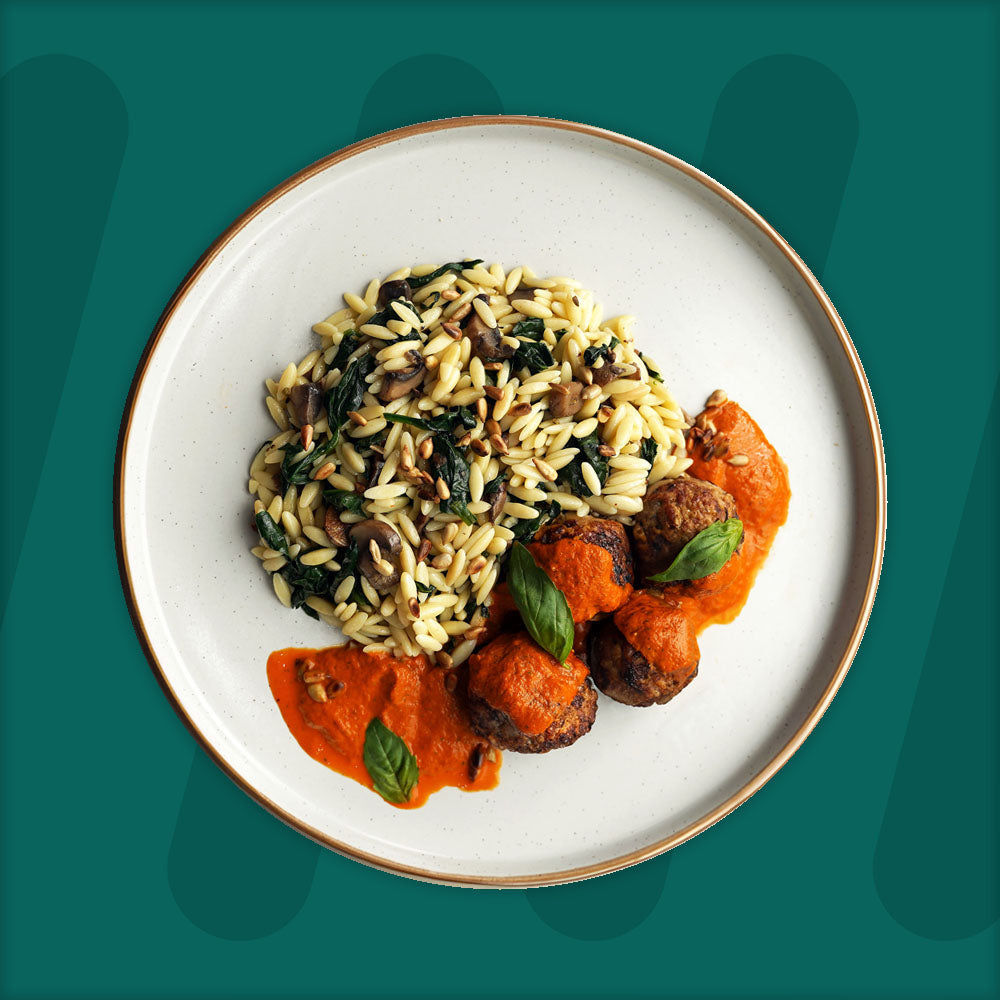 A plate featuring flavorful meatballs topped with a rich red pepper sauce and garnished with fresh basil leaves, accompanied by a serving of orzo pasta mixed with sautéed spinach, mushrooms, and toasted seeds, set against a green background.