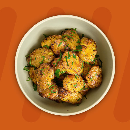 A bowl of golden roasted baby potatoes, seasoned with herbs and garnished with fresh parsley, displayed against a vibrant orange background