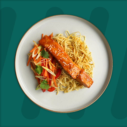 This delightful plate features a glazed salmon fillet, coated with a shiny teriyaki sauce and topped with sesame seeds for added texture and flavor. It rests atop a bed of golden egg noodles, perfectly cooked to retain a slight bite. On the side, a refreshing salad of julienned carrots, red bell peppers, and green onions is dressed lightly, bringing a crunchy and vibrant contrast. The meal is elegantly presented on a minimalist plate, set against a deep green background, emphasizing the dish's colorfulness